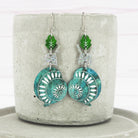 A pair of acrylic snail dangle earrings with green and blue patterns, attached to silver titanium ear wires, displayed on a concrete block.