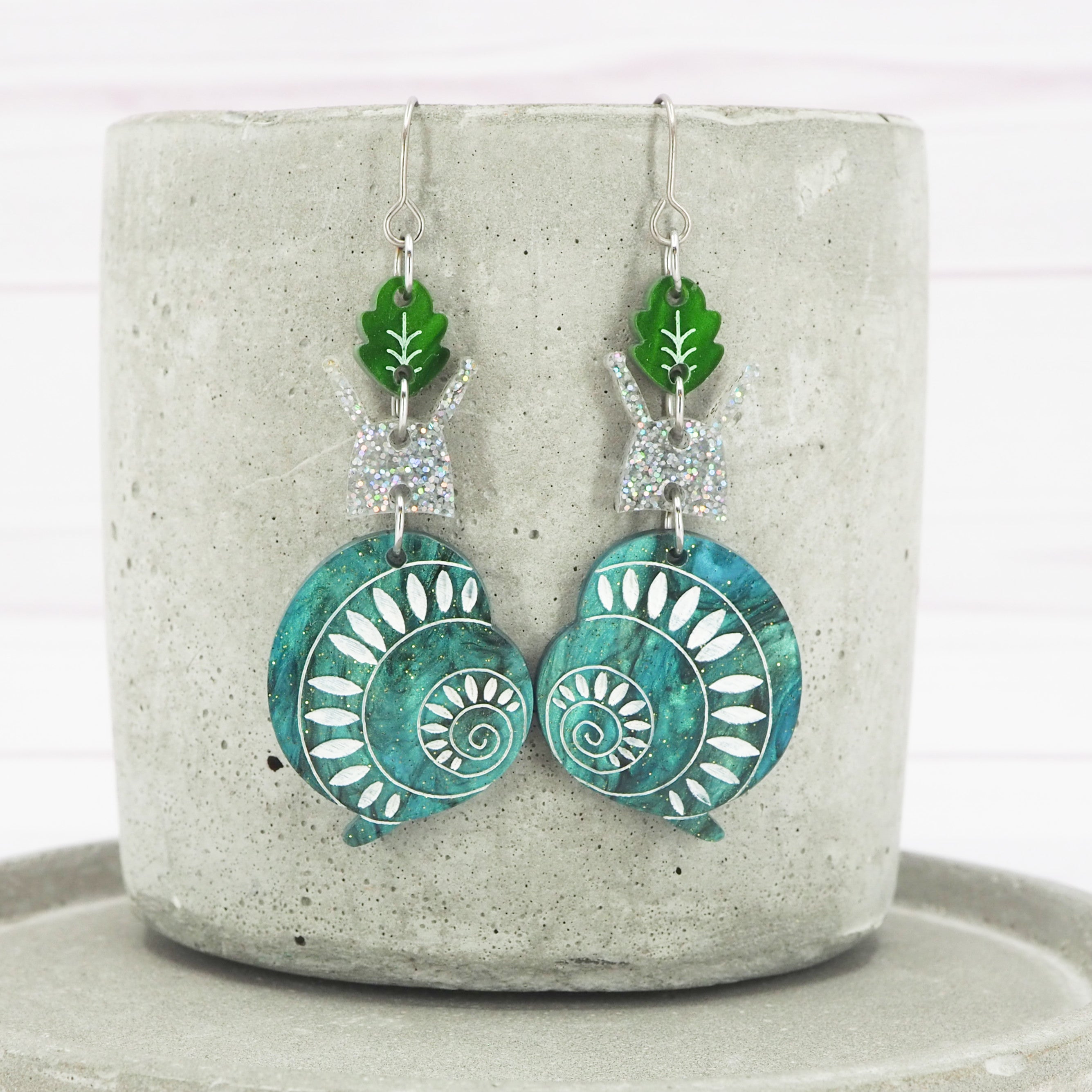 A pair of acrylic snail dangle earrings with green and blue patterns, attached to silver titanium ear wires, displayed on a concrete block.