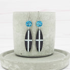 A pair of glossy black acrylic surfboard dangle earrings with St Piran flag design, displayed on a grey ceramic holder.