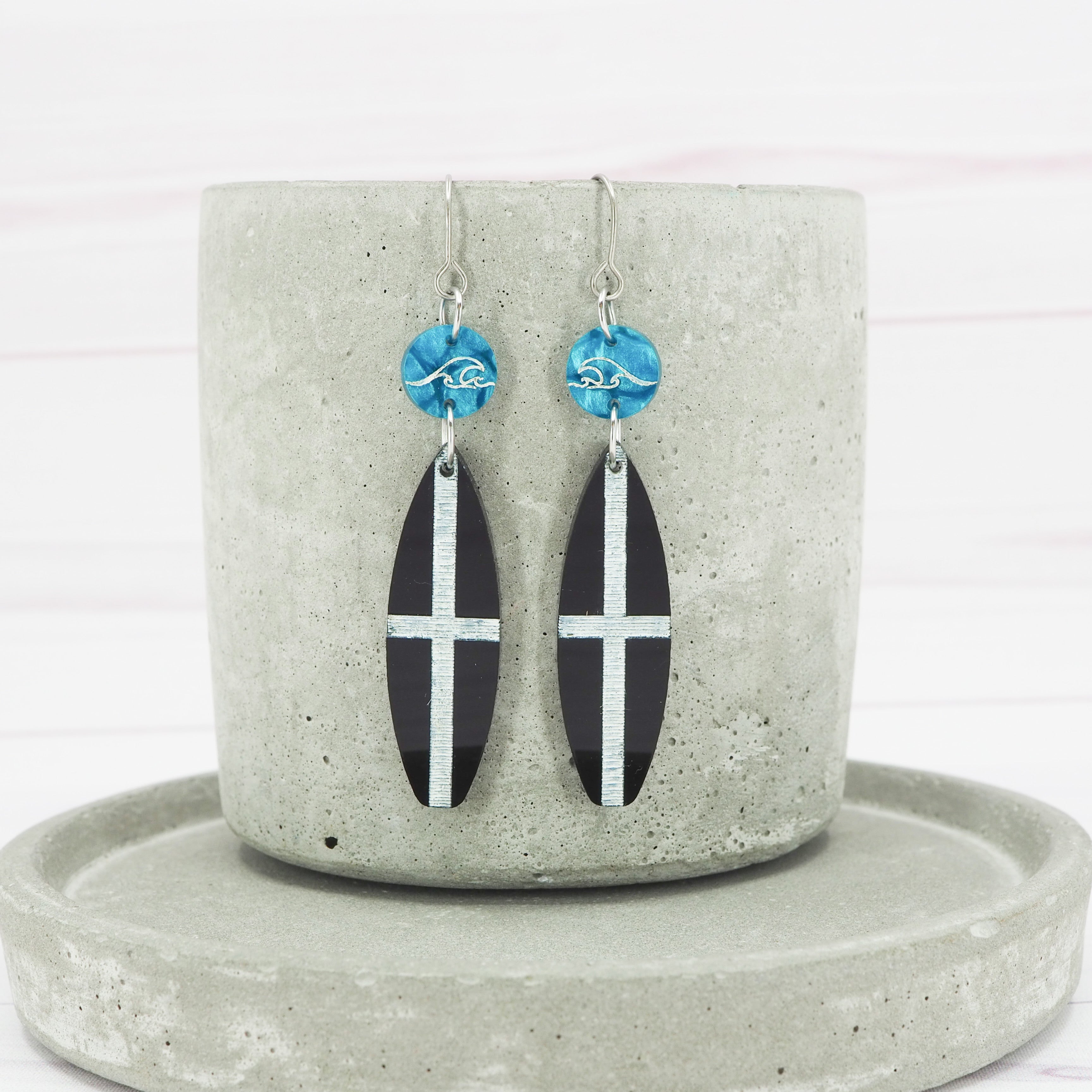 A pair of glossy black acrylic surfboard dangle earrings with St Piran flag design, displayed on a grey ceramic holder.