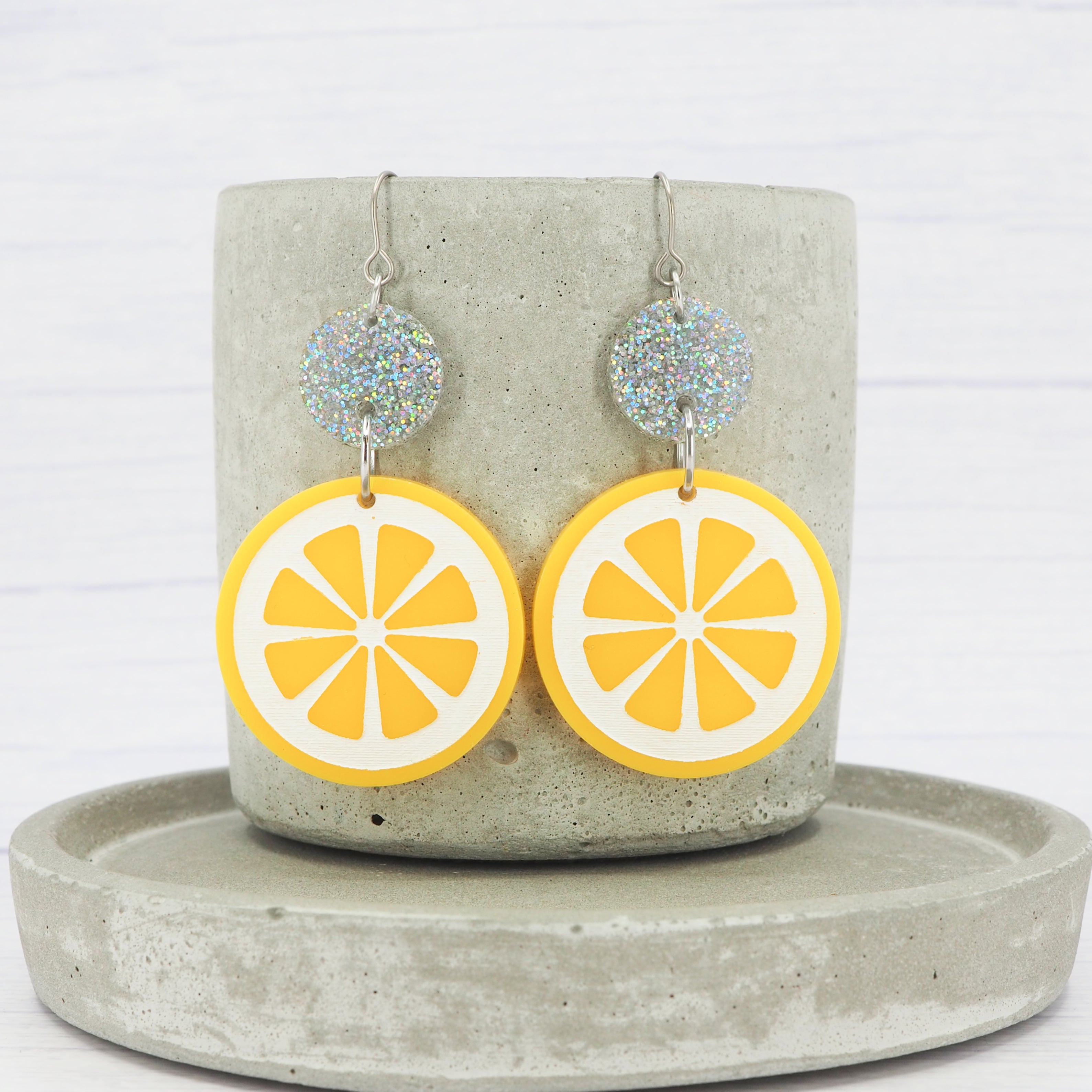 A pair of glossy yellow lemon-shaped dangle earrings with titanium ear wires, displayed on a gray stand.